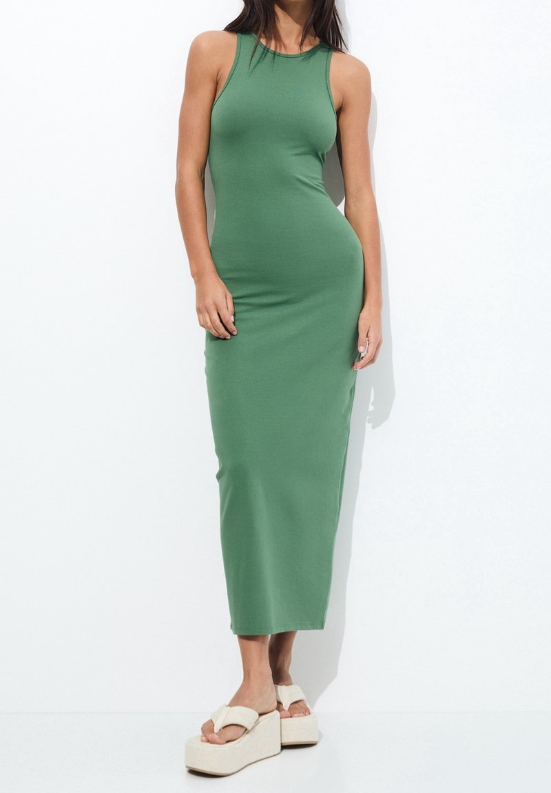 Femme portant une robe midi verte ajustée sans manches associée à des sandales à plateforme beige, debout sur un fond blanc uni.