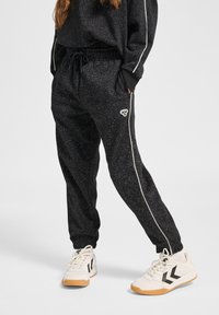 Schwarze Jogginghosen mit einem gesprenkelten Muster, elastischem Bund und weißen Seitenstreifen. Kombiniert mit weißen Sneakers mit schwarzen Akzenten.