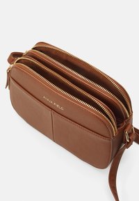 Brun læder crossbody taske med tre lynlåslommer, guldfarvede lynlåsdetaljer og et præget logo på forsiden.