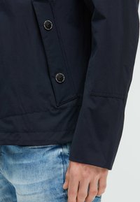 Granatowa kurtka wykonana z teksturowanej tkaniny, z zapięciem na guziki oraz dopasowanym krojem z wyraźnymi ramionami i prostym dołem.