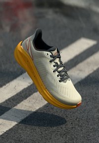 Hellgrauer Sportschuh mit strukturiertem Obermaterial, gelber Sohle, grauen Schnürsenkeln und orangefarbenen Akzenten. In der Luft vor einem dunklen Hintergrund positioniert.