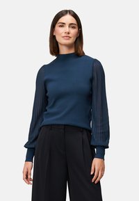 Marineblå sweater med høj hals, der har en fast strikket krop og gennemsigtige, plisserede lange ærmer. Sammen med skræddersyede sorte bukser.