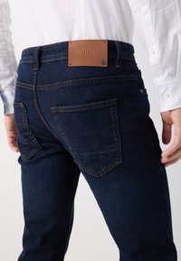 Jeans in denim blu scuro con una toppa di pelle del marchio sul retro, caratterizzati da tasche posteriori e dettagli di cuciture a contrasto.