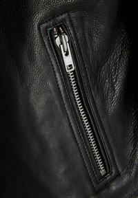 Black leather jacket detail featuring a visible YKK zipper and textured surface. The zipper is silver with a contrasting fabric lining.