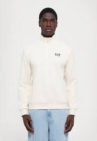 Kremfarget genser med høy hals, halv glidelås og svart logo på brystet. Mykt materiale og avslappet passform. Kombinert med lyseblå jeans.
