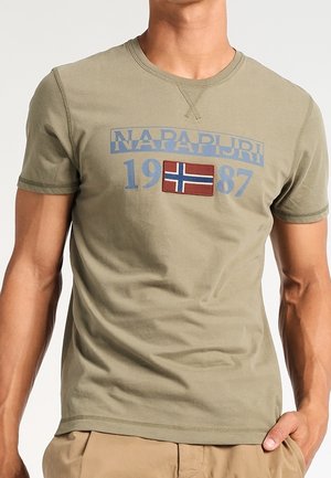 Homme portant un t-shirt Napapijri vert olive avec "1987" et un patch du drapeau norvégien sur la poitrine, assorti à un pantalon beige.