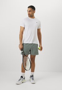Hvit treningst-skjorte, grå shorts med logo, holder en tennisracket og ball, iført hvite og marineblå sportsko, står på en nøytral bakgrunn.