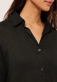 Camicia di lino nera con colletto, chiusura anteriore con bottoni e texture visibile. Presenta una vestibilità ampia e un tessuto morbido.