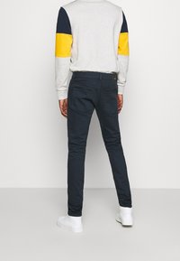 Hombre vistiendo pantalones ajustados azul marino, un sudadera ligera gris claro con bloques de color amarillos y azul marino en las mangas, y zapatillas blancas, de pie contra una pared blanca.