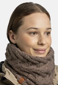 Knitted brown scarf, chunky texture with a braided pattern, featuring a leather tag. Worn around the neck, part of a neutral outerwear outfit.