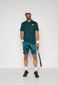 Herren-Tennis-Outfit mit einem dunkelgrünen, strukturierten Poloshirt und passenden Shorts. Akzente bilden ein weißes Logo und schwarze Sportschuhe.