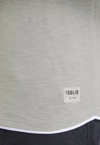 Tricou gri deschis din material moale, cu o textură subtilă și tiv alb. Prezintă o etichetă de marcă rectangulară cu text negru.