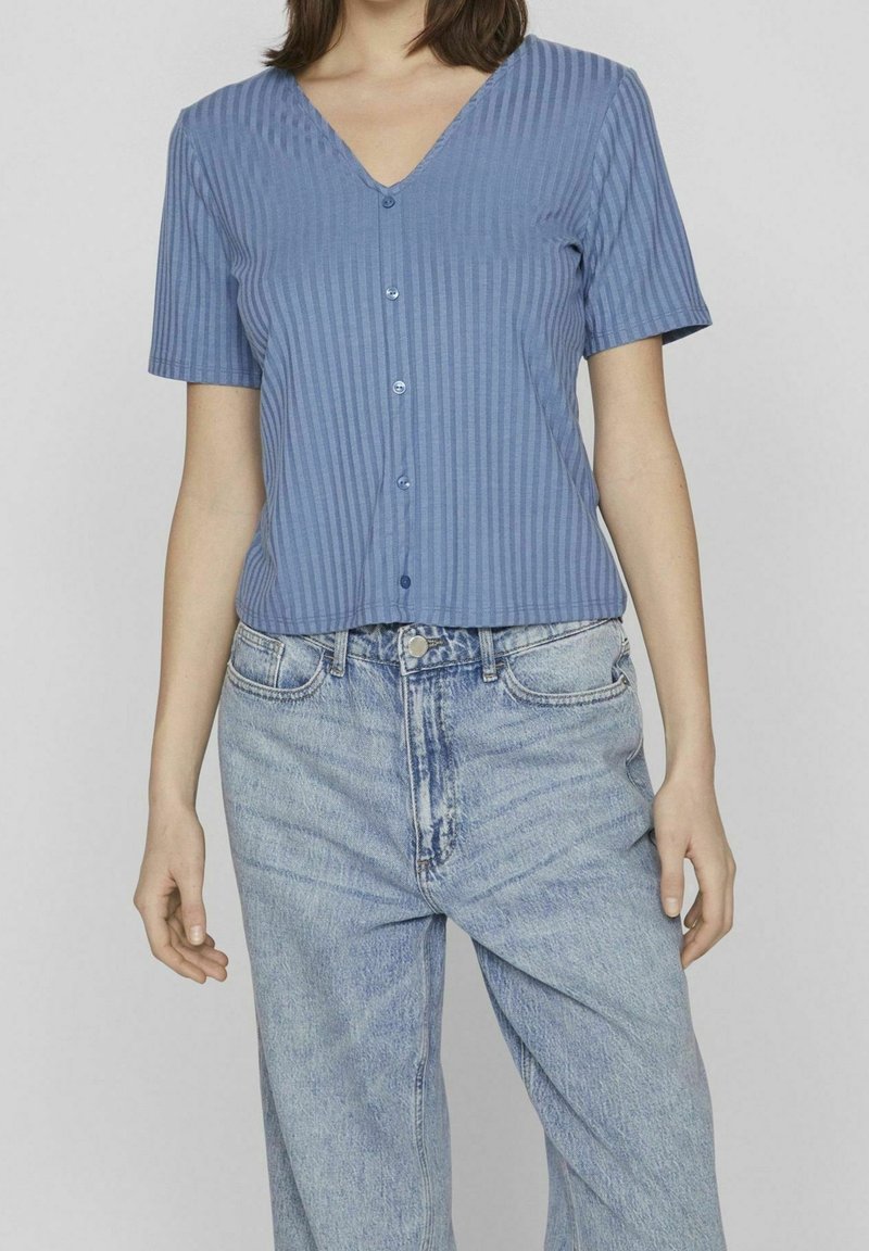 Žena oblečená v krátkém rukávu modré pruhované košili na knoflíky a světlých modrých džínách s vysokým pasem, stojící na jednobarevném pozadí.