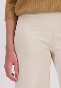De hand van een vrouw rust naast haar, ze draagt beige broek en een bruine geribbelde trui waarvan de mouw zichtbaar is boven de pols.