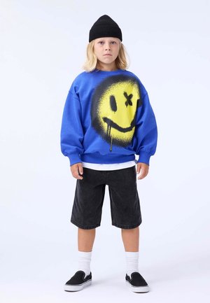 Felpa blu con una grande grafica di un volto sorridente giallo, pantaloni corti neri, calze bianche e sneakers nere, completato con un berretto nero.