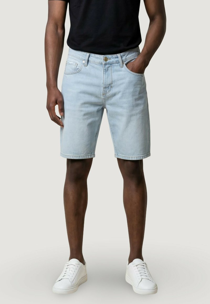 Mężczyzna w jasnoniebieskich krótkich spodenkach dżinsowych, czarnej koszulce i białych trampkach, stojący z jedną ręką w kieszeni na jednolitym tle.