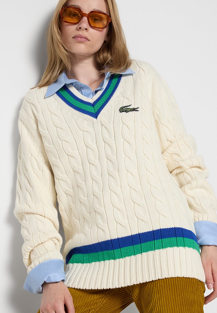 Maglione in crema a maglia cable-knit con scollo a V, caratterizzato da un colletto a righe verdi e blu. Logo Lacoste vicino al collo. Camicia azzurra leggera sotto.