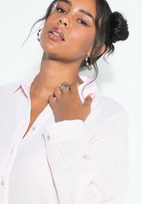Camicia button-up rosa chiaro, con texture e colletto morbido. Sono indossati orecchini in argento e un anello elaborato, insieme a smalto per unghie rosa chiaro.