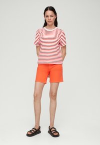 Rood en wit gestreept T-shirt met een ronde hals, gecombineerd met fel oranje shorts. Model draagt zwarte sandalen. Simpele, casual outfit.