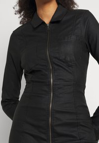 Svart figursydd jumpsuit gjord av stretchigt material, med dragkedja fram, krage och två bröstfickor. Texturerad yta med synliga sömmar.