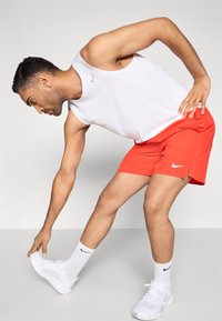 Λευκό αμάνικο μπλουζάκι με το λογότυπο της Nike, κόκκινο αθλητικό σορτς, λευκά αθλητικά παπούτσια και κάλτσες με το έμβλημα της Nike· σε θέση να πιάνει ένα παπούτσι.