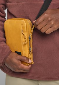 Geel ritstas met een gladde textuur, voorzien van een zwarte accent en een afneembare ritshanger, vastgehouden door een persoon in een donker kastanjebruine fleece.