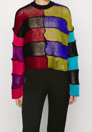 Person iført en løsstrikket sweater med horisontale farveblokke i rød, sort, lilla, olivengrøn, blå og turkis med sort kant og teksturerede sorte bukser.