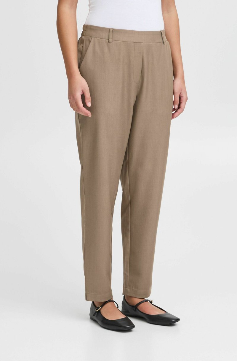 Pantalon beige ajusté avec une texture lisse, présentant un devant plat, des poches latérales et une coupe décontractée, associé à des chaussures plates noires.