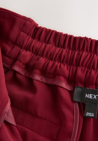 Bordeauxfarvet elastisk taljebånd med rynket tekstur, glat inderfor, mærke med teksten "NEXT," fremstillet i Vietnam, blødt stoffet detaljer synlige.