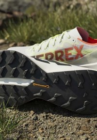 Weiße und orangefarbene Trailrunning-Schuhe mit Continental-Gummisohlen liegen auf Kies mit Gras im Hintergrund.