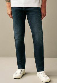 Jeans de denim albastru închis cu o croială dreaptă, prezentând o textură subtilă și detalii clasice de cusătură, asortate cu adidași albi.