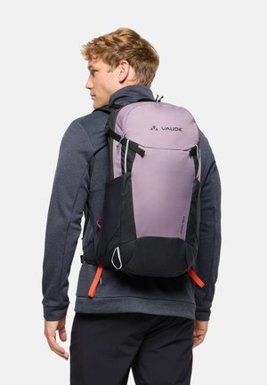 Giovane uomo che indossa uno zaino Vaude viola e nero con cinturini rossi, vestito con una giacca scura texturizzata e pantaloni neri, volto verso sinistra.