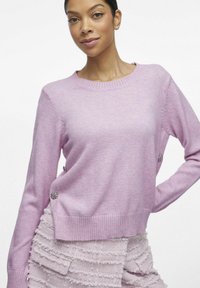 Fioletowy sweter z okrągłym dekoltem, długimi rękawami i ozdobami po bokach. Miękka tekstura i prążkowany dół tworzą wygodny wygląd.