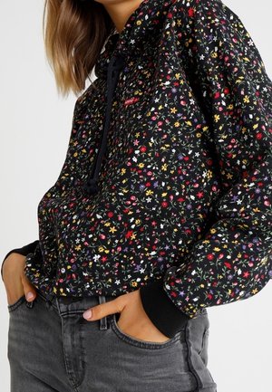 Femme portant un sweat à capuche noir à fleurs avec de petites fleurs colorées, des poignets noirs et un jean gris foncé, mains dans les poches.