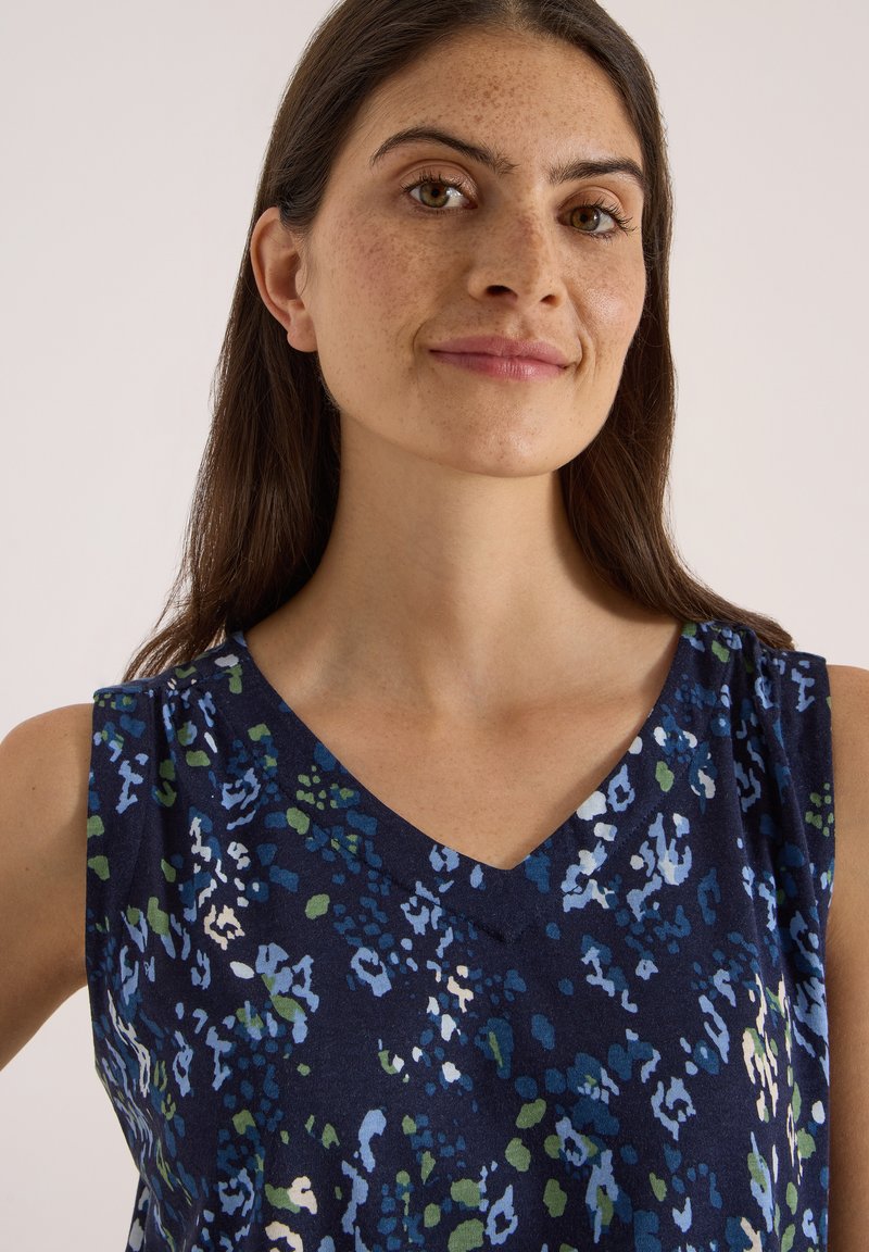Femme aux longs cheveux bruns et taches de rousseur portant une robe bleu marine sans manches avec un motif floral abstrait vert, blanc et bleu clair.