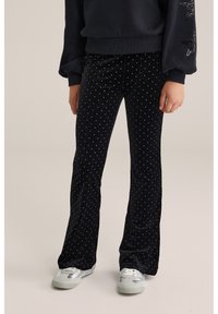 Črne žametne hlače s širokimi nogami in belimi pikami, ki imajo mehak teksturo in prilegajoč dizajn. V kombinaciji s srebrnimi tenisicami.