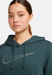 Tyrkysová mikina s kapucí a stahovací šňůrkou, na přední straně s vyšitým stříbrným logem Nike. Měkká textura a volný střih.