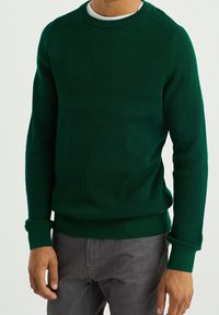 Donkergroene geribbelde trui met een ronde hals en lange mouwen, voorzien van een textuurstof en aansluitende manchetten, gecombineerd met grijze broek.