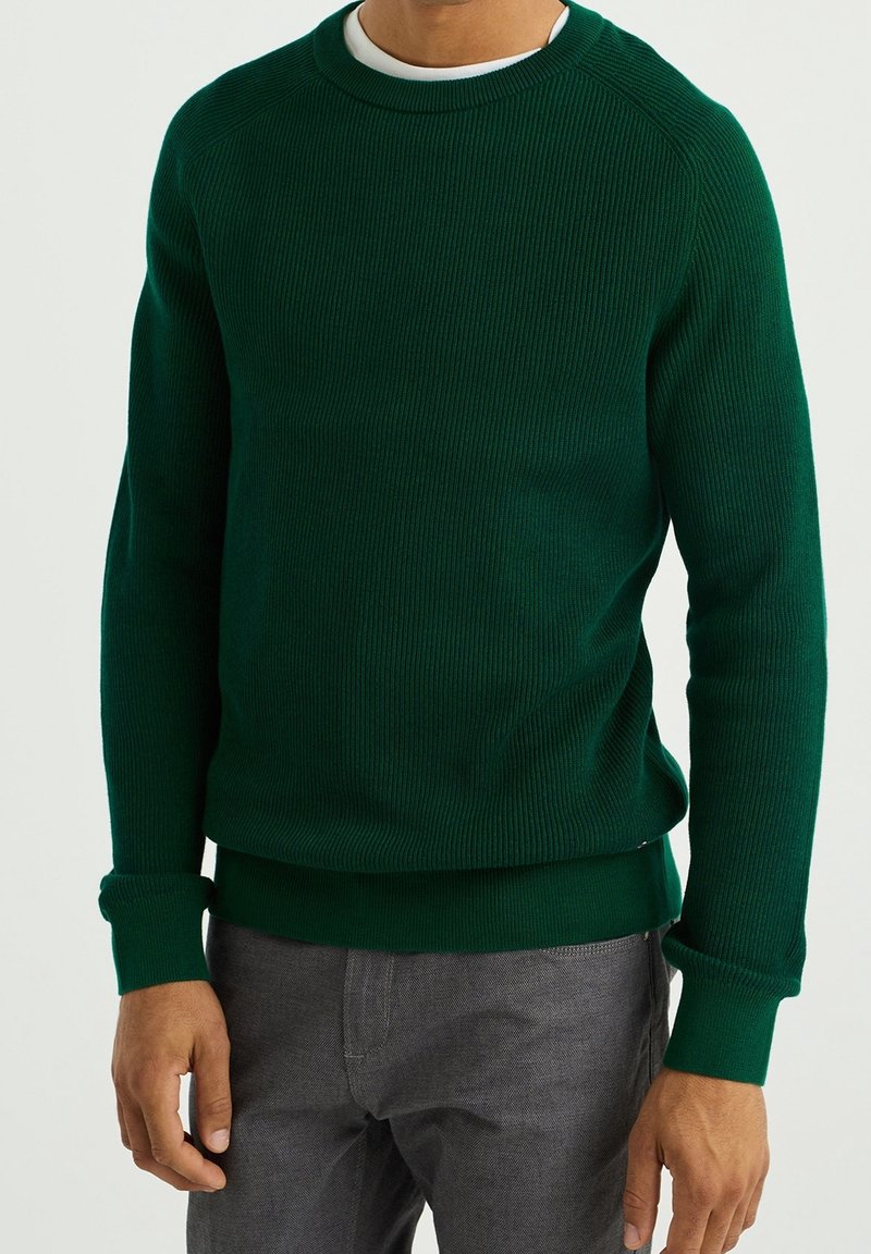 Donkergroene geribbelde trui met een ronde hals en lange mouwen, voorzien van een textuurstof en aansluitende manchetten, gecombineerd met grijze broek.