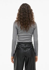 Persona con lunghi capelli ricci, che indossa un top grigio a maniche lunghe e pantaloni in pelle neri a vita alta, girata di spalle su uno sfondo bianco.