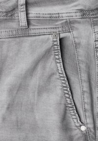 Gros plan sur une poche de jean en denim gris clair avec coutures visibles, passants de ceinture et rivets métalliques.