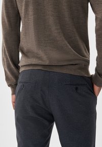 Braun fein gestrickter Pullover und dunkelblaue Hose mit strukturierter Oberfläche; die Hose verfügt über zwei Gesäßtaschen mit Knöpfen.