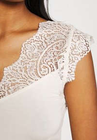 Biała bluzka z koronkowymi rękawami w kształcie motyla, zdobiona skomplikowanymi wzorami kwiatowymi oraz gładkim, dopasowanym krojem. Miękka tekstura z okrągłym dekoltem.