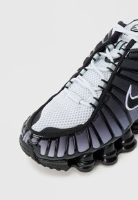 Tenisky Nike s bílým síťovinovým svrškem přecházejícím do tmavě šedé. Silná, texturovaná podrážka s černými detaily a ozdobnými vzory.