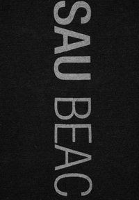 Czarna tkanina z wyraźnym szarym napisem "SAU BEAC" w pionowym układzie; miękka tekstura z gładkim wykończeniem.