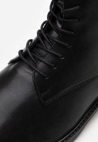 Botín de cuero negro con una textura suave, puntera redonda, costuras reforzadas y cordones negros contrastantes con ojales de metal.