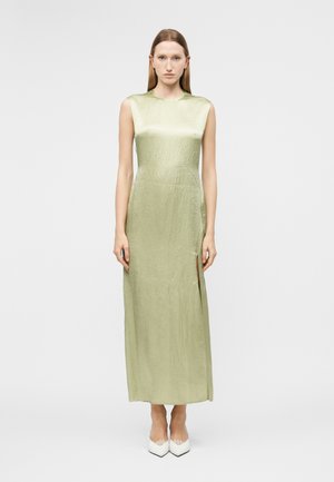 Kvinde med glat rødt hår iført en ærmeløs, lysegrøn, tekstureret kjole til anklerne med en sideslid og hvide spidse hæle, stående vendt fremad.