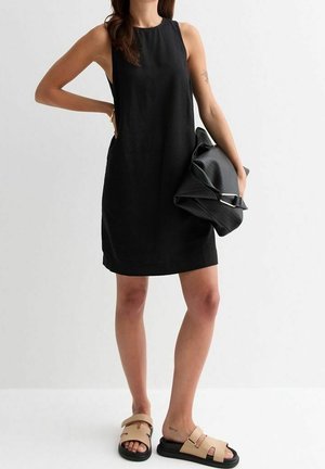 Kobieta w czarnej sukience bez rękawów i beżowych sandałach, trzymająca dużą czarną teksturowaną kopertówkę na białym tle.