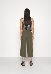 Woman with long dark hair wearing a black floral sleeveless top, olive wide-leg cropped pants, and black sandals, standing facing a white wall.