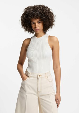Kobieta o kręconych włosach, ubrana w białą, bezrękawnikową, prążkowaną bluzkę i beżowe spodnie z wysokim stanem, stoi na tle białego tła z jedną ręką w kieszeni.